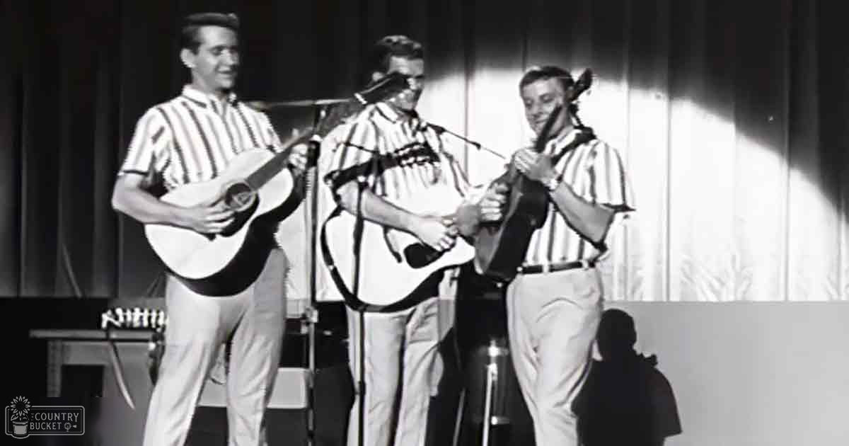 Musical performance by three adult males in an indoor venue with microphones and a curtain backdrop; evening lighting and grayscale photography.