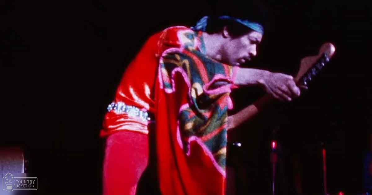 A middle-aged male musician in a vibrant red, patterned robe performs on stage at an indoor concert with stage lighting focused on him. A blue headband is worn.