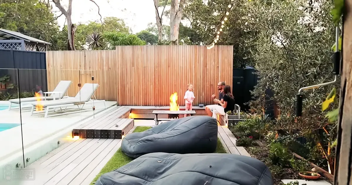 A backyard patio area with lounge chairs, string lights above a fire pit, surrounded by greenery and trees on a clear day.