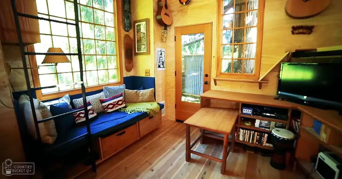 A cozy indoor space with wooden furniture, books, and a guitar hanging above the desk, large windows showing greenery outside, likely spring or summer.