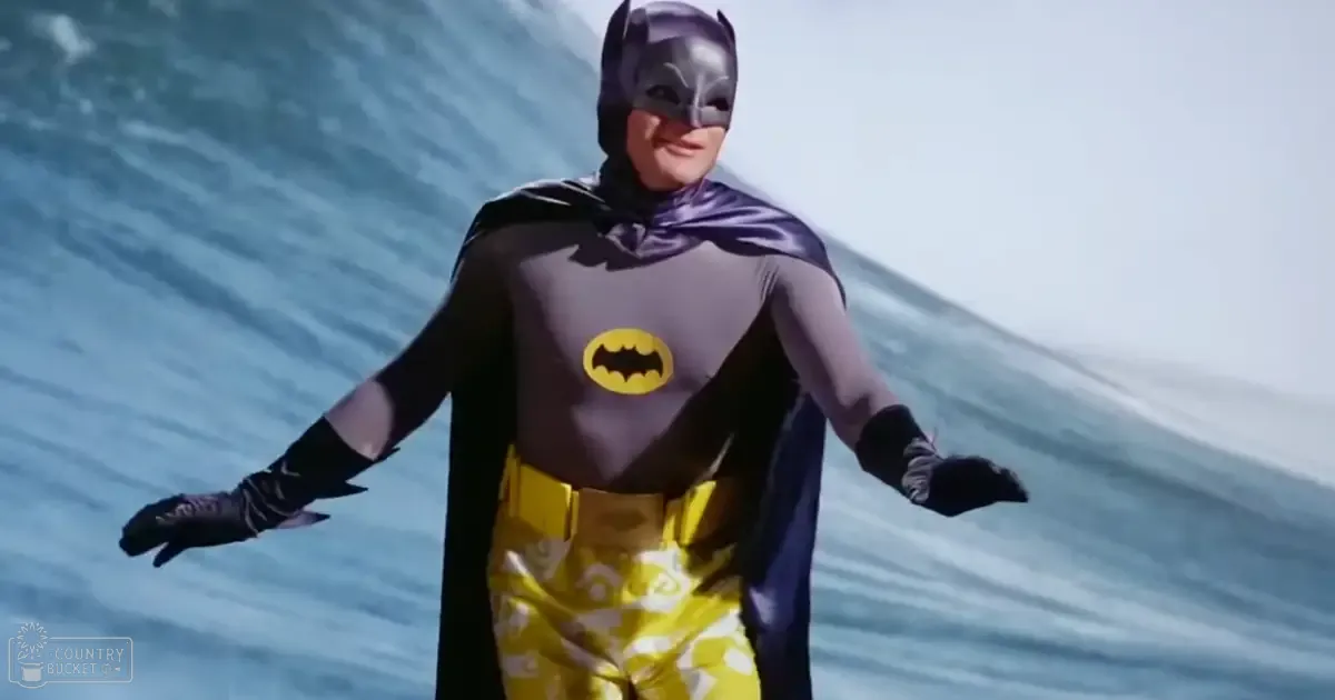 A person dressed in a Batman costume stands against an ocean backdrop under bright daylight.