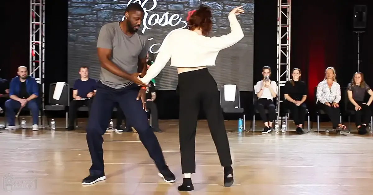 A dance performance scene with a male and female dancer in an indoor setting, surrounded by spectators. The backdrop features 'Rose' written on it.
