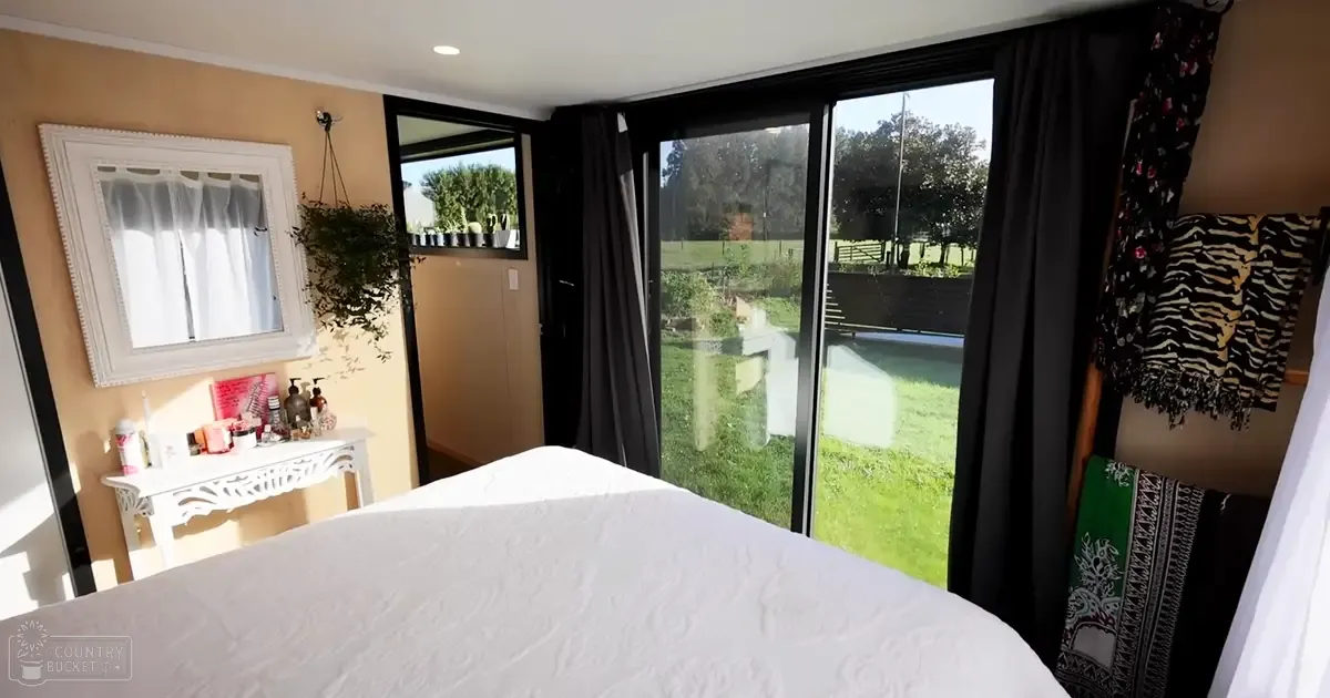 A bedroom scene with a white quilted blanket, zebra-patterned throw, black curtains, sliding glass doors leading to an outdoor area with grass and trees visible. The light suggests daytime during spring or summer.