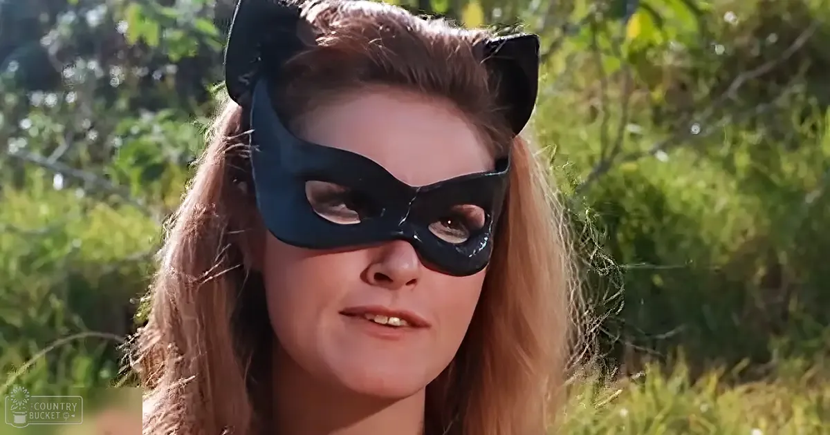 A young adult female wearing a black mask with cat-like ears and eye cutouts stands in an outdoor setting with greenery, under natural daylight.