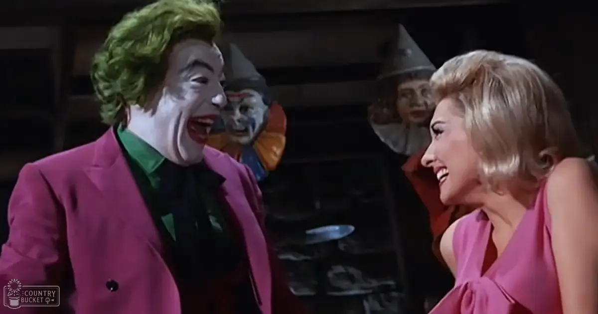 A theatrical or film setting with characters in vintage attire from the mid-20th century; adult male on left in bright pink suit jacket and green hair, adult female on right in sleeveless pink dress with blonde bob hairstyle.