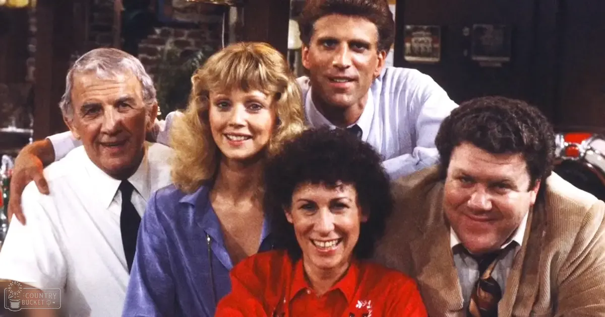 A group photo from a television show set in a living room with brick fireplace, framed picture, red car partially visible behind the group.