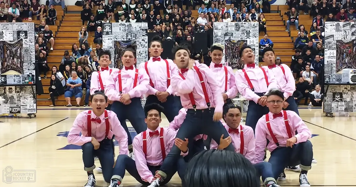 A group performance by young adult males in a gymnasium with pink dress shirts, red suspenders, black bow ties, dark jeans or pants, and black sneakers with white soles.