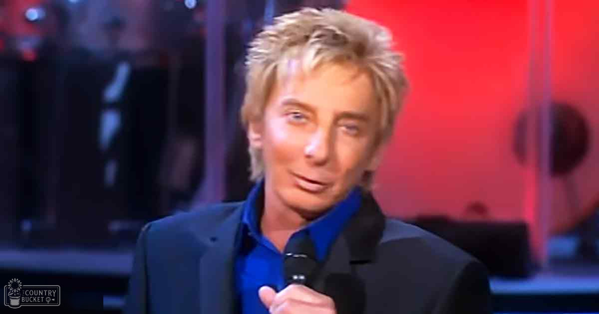 A middle-aged man wearing a dark blazer and holding a microphone on an indoor stage with red lighting.