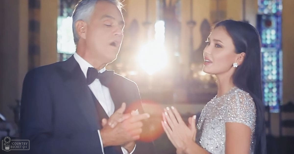 A middle-aged man in a black tuxedo holds something red while standing next to a young woman in a silver sequined dress at an indoor formal event with stained glass and chandelier lighting.