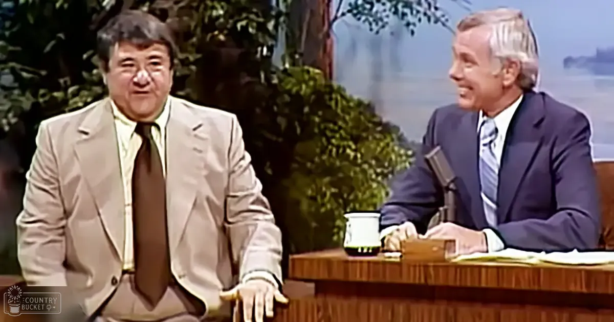 A middle-aged male talk show host in a beige suit with white shirt and dark tie, seated at a wooden desk with another older adult male guest wearing a dark blue suit and light blue striped tie. The setting is a television studio with blurred outdoor scenery and even lighting.
