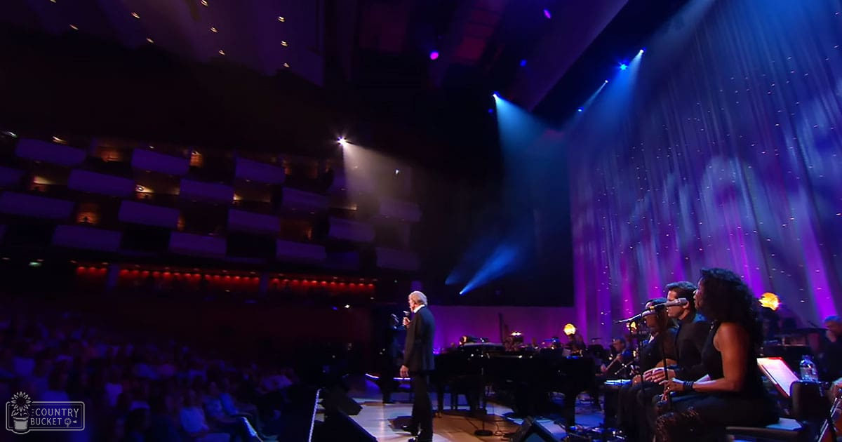 A concert scene in a theater, with an older adult male performer in a dark suit holding a microphone on stage alongside a young adult female singer in a black dress. Musicians with instruments are seated behind them under dramatic lighting.