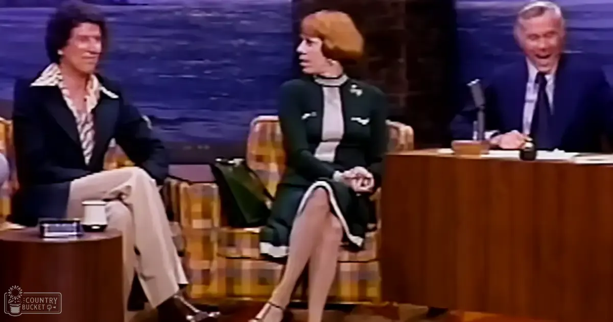 A talk show set with three individuals: an adult male host in a dark blazer over a patterned shirt and light trousers, an adult female guest in a black dress with white trim at the hemline and sleeves paired with high-heeled shoes, and another adult male guest dressed in formal attire seated behind a desk. The setting includes studio furniture like chairs and a wooden desk with a microphone, under artificial lighting.