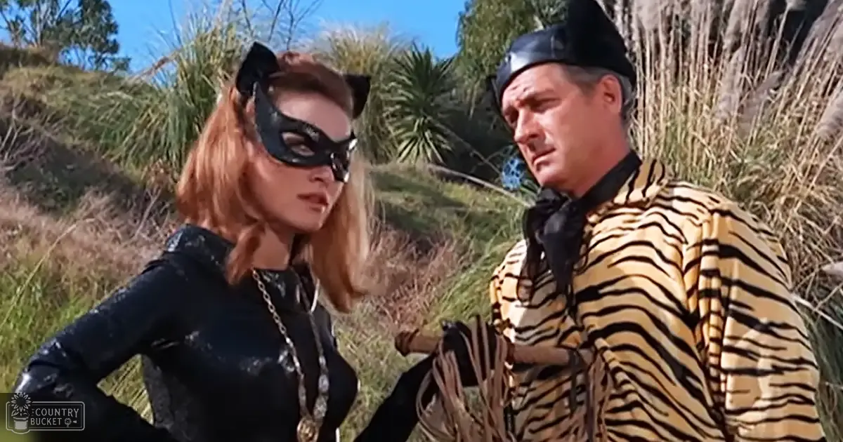 Movie or TV show scene outdoors with adult female in a cat-ear costume and male in tiger-striped jacket on hillside, spring or summer day.
