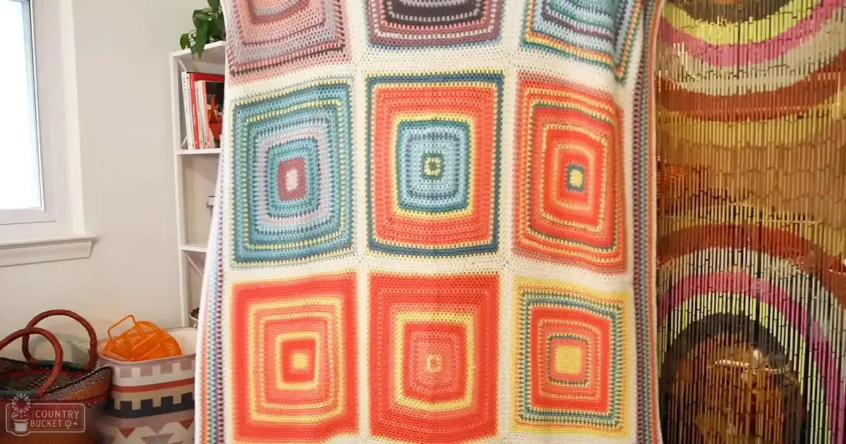 A colorful crocheted blanket with a grid pattern of squares in various colors indoors near bookshelves and natural light coming through a window.