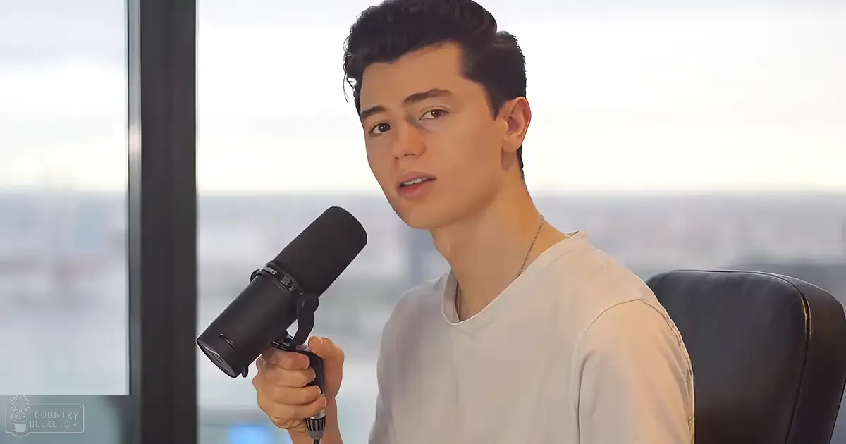 A young adult male wearing a relaxed fit white t-shirt with a modern casual style, holding a black microphone in an indoor setting with large windows showing an overcast cityscape.