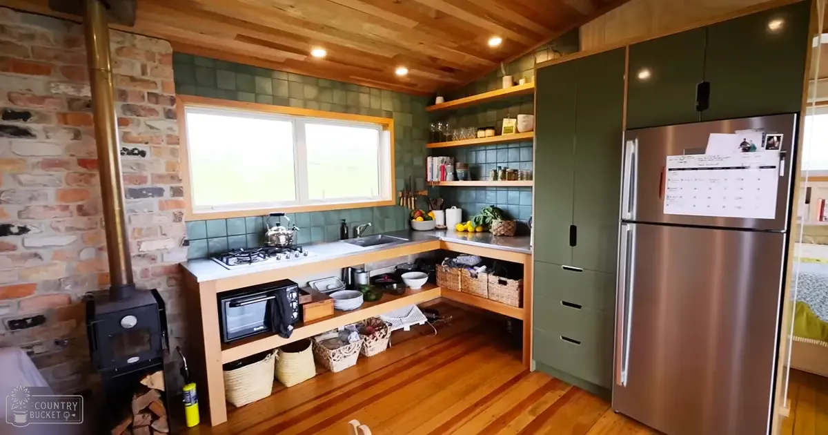 A cozy home interior with a stainless steel refrigerator, gas stove with kettle, wooden shelves holding books and jars, open shelving containing baskets, a brick wall wood-burning stove, two windows allowing natural light into the room; indoors in what appears to be a home environment.