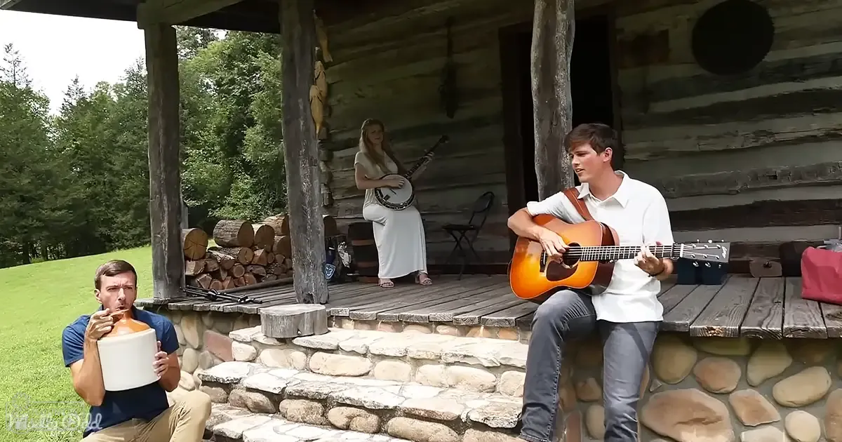 Musical performance outdoors on a rustic log cabin porch with green grass and trees in the background, late spring or summer day.