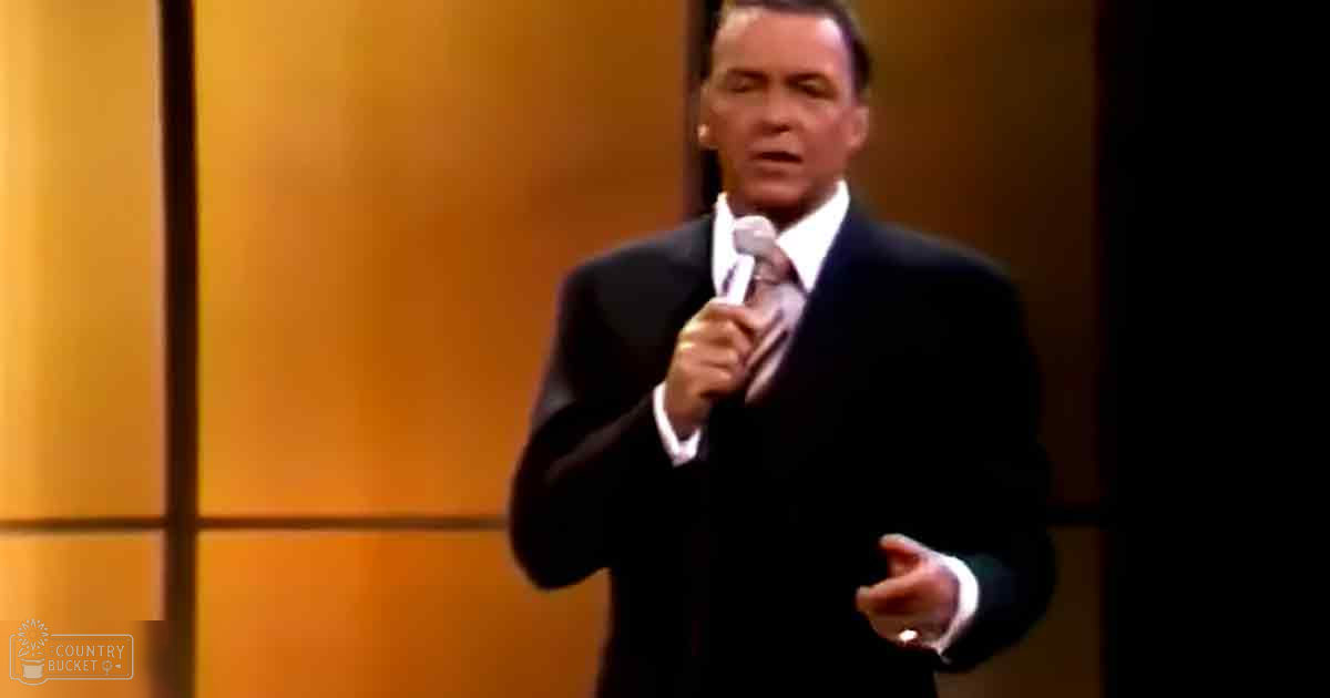 Adult male giving a speech or presentation in an indoor setting, wearing a dark suit jacket, white shirt, and patterned tie.