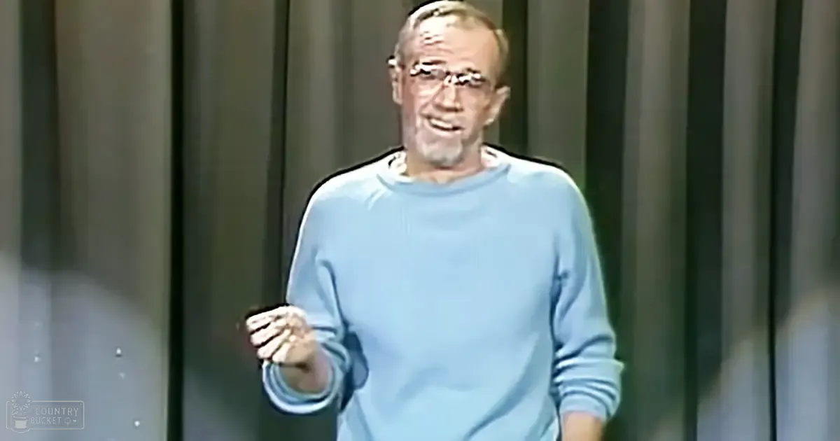 An older adult male in a light blue long-sleeved sweater, holding a microphone indoors with curtains or blinds in the background.