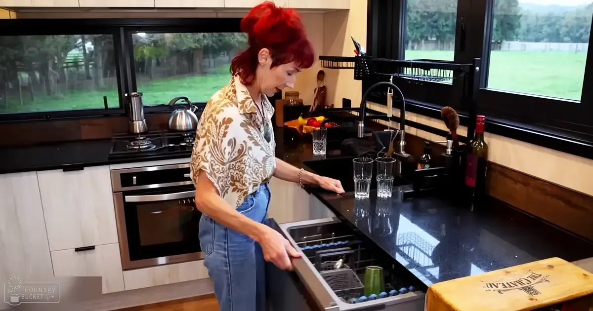 A middle-aged woman wearing a floral patterned blouse and blue jeans in her modern kitchen setting with black countertops, stainless steel oven, sink area, wooden cutting board, bottle of wine, fruit basket, and dish drying rack.