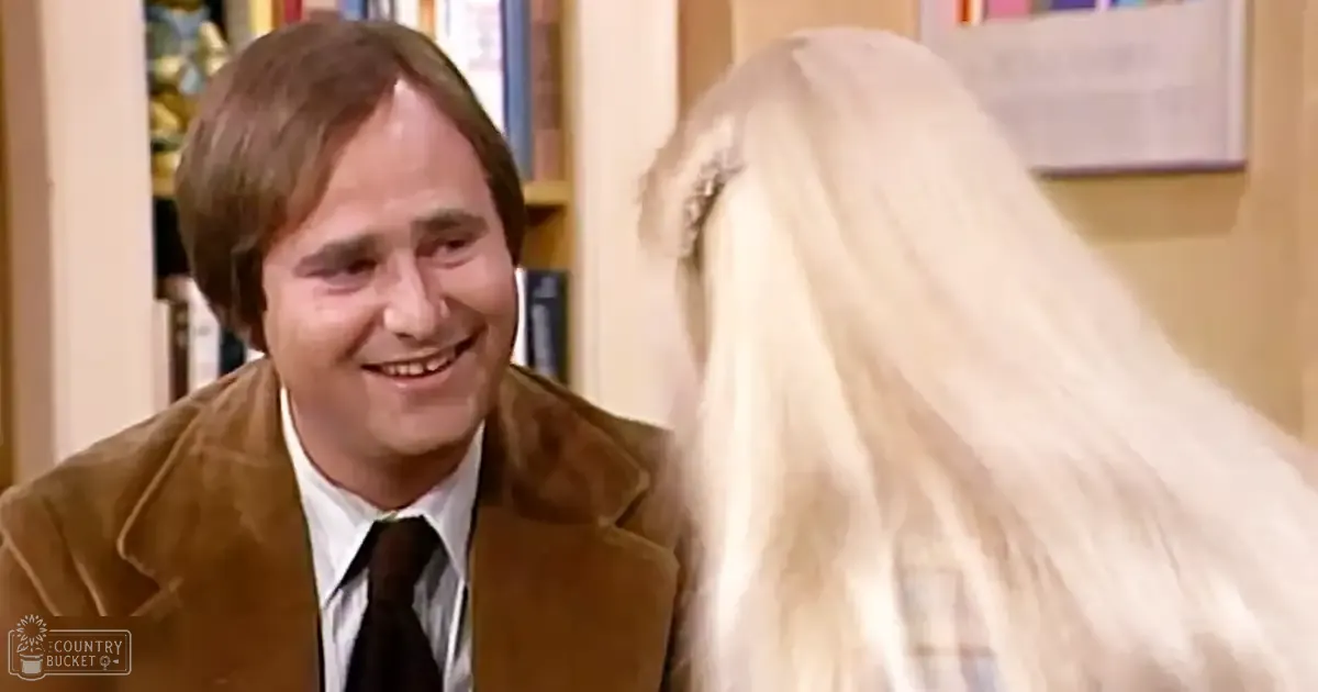 An adult male and female in a room with bookshelves. The man wears a brown suede jacket over a white collared shirt and dark tie, suggesting mid-20th-century formal style; the woman has long blonde hair under a white veil.