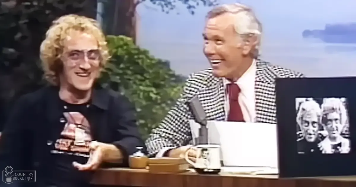 A talk show host and guest in a late 20th-century studio setting with scenic backdrops; adults wearing casual to formal attire, surrounded by typical furniture items.