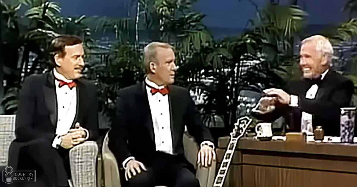 Three men in 1960s or 1970s tuxedos on a talk show set with tropical-themed decor, indoor studio lighting.