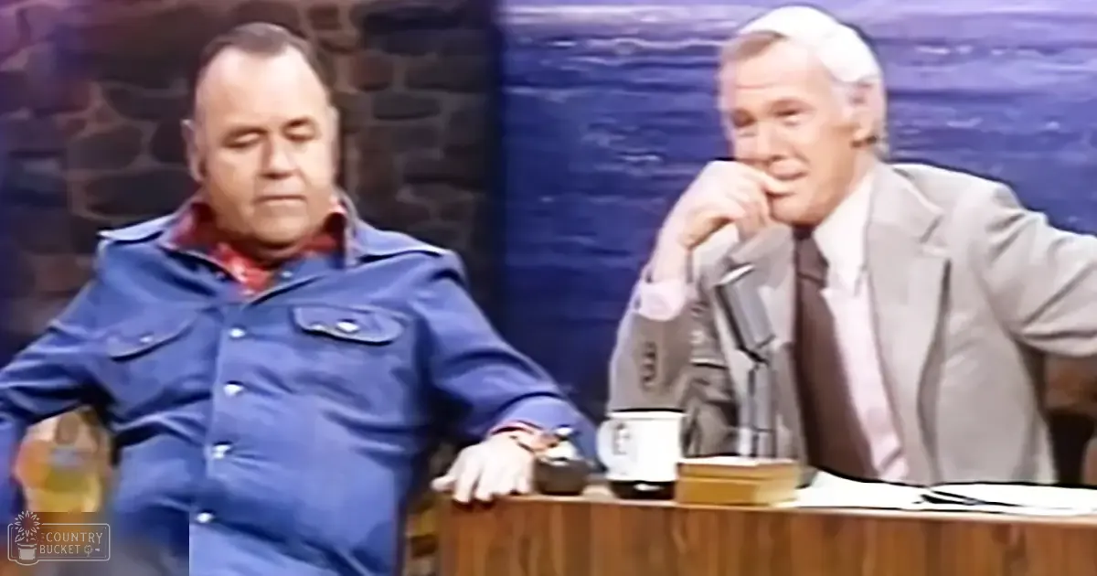 Two men in a studio setting, one middle-aged wearing a blue button-up with red plaid pattern under a dark jacket, seated at a wooden desk with a white mug and small box. The other is an older adult male in a light gray suit and dark tie, also seated at the desk.