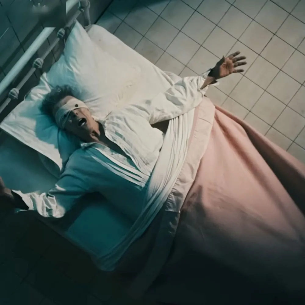 An older adult male wearing a white shirt, dark green or blue undergarments, and pink blanket in a metal-framed bed with tiled surroundings, suggesting an institutional setting.