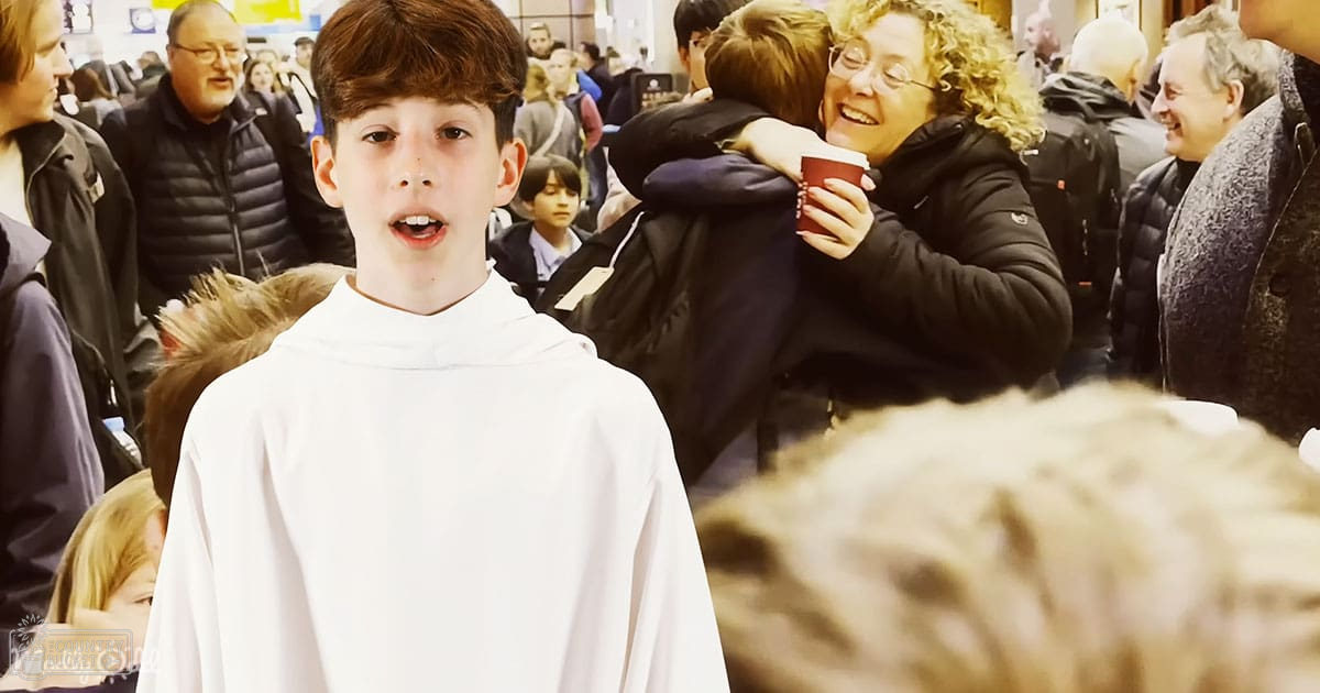 A young boy in white robes stands indoors amidst a crowd dressed for cooler weather during the daytime.