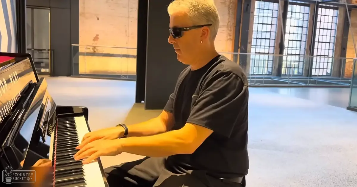 A middle-aged man wearing a black t-shirt and dark gray pants plays a grand piano in a modern industrial-style interior with large windows allowing natural light to enter, suggesting daytime.