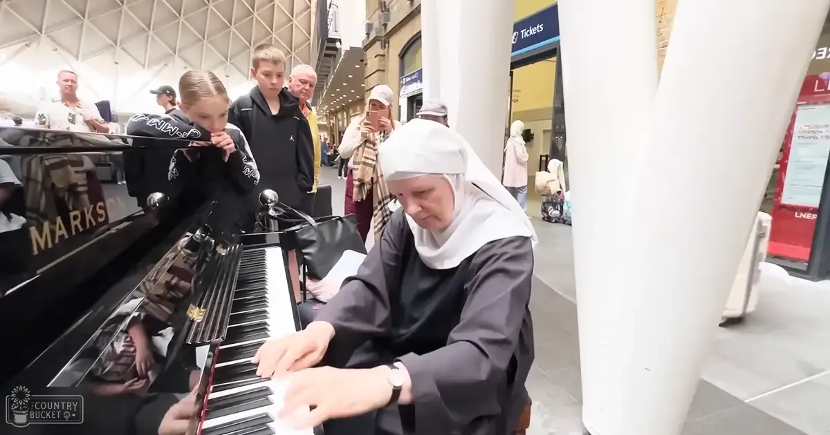 A nun playing piano with a young girl and a teenager at a train station, surrounded by other passengers during daytime.