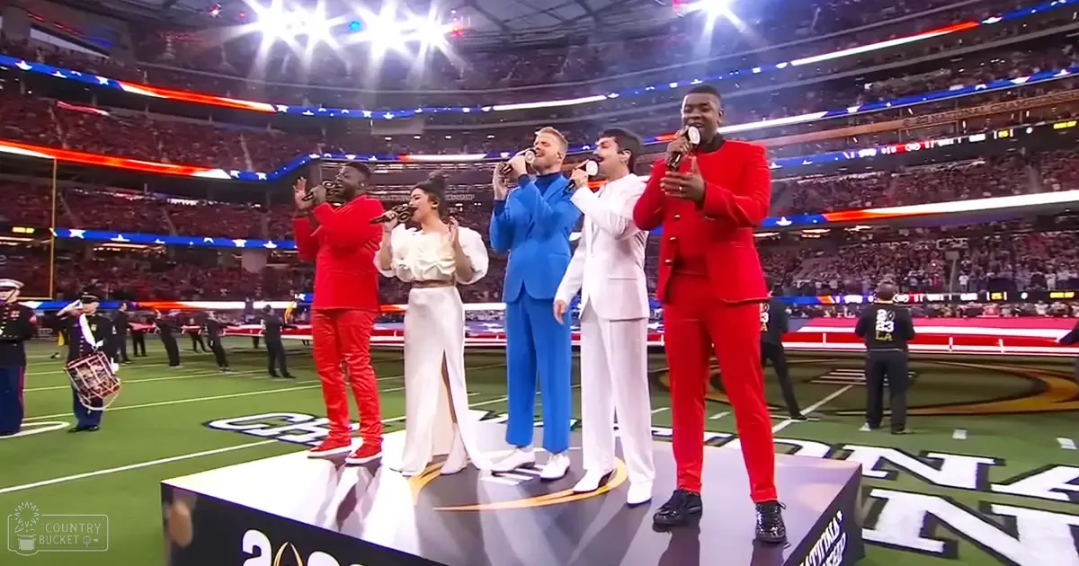A group performing at an event in a large indoor stadium, illuminated by bright lights under evening skies; spectators fill the stands.