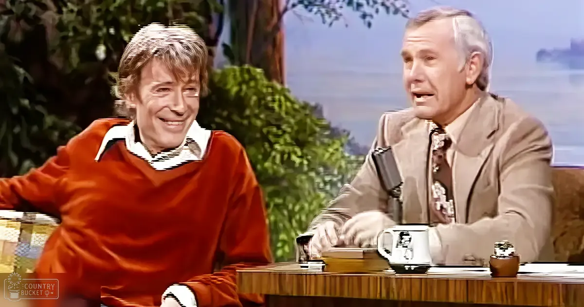 Two middle-aged men on a talk show set, wearing casual business attire. A wooden desk with everyday items and a backdrop of trees and sky.