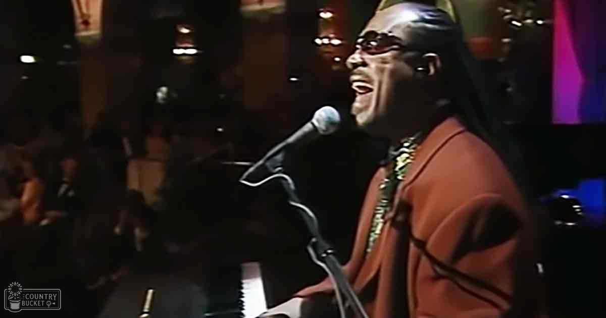 An adult male singer in a red jacket with black accents and a gold chain, singing into a microphone at an indoor concert or performance setting.