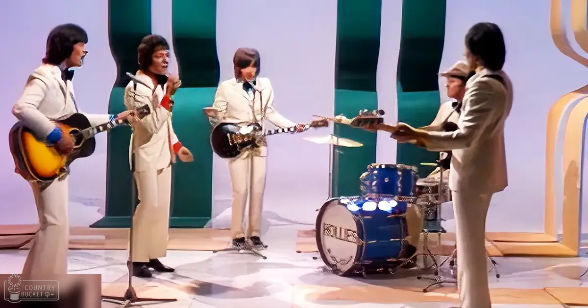 A musical performance with four adult males in white suits and one drummer in a beige suit, set in a studio environment.