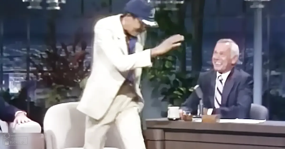 A man in a tailored white suit jacket and beige pants dances on a talk show set, while another adult male sits wearing a dark formal suit with a striped tie. The setting includes a desk with a microphone and urban views through large windows.