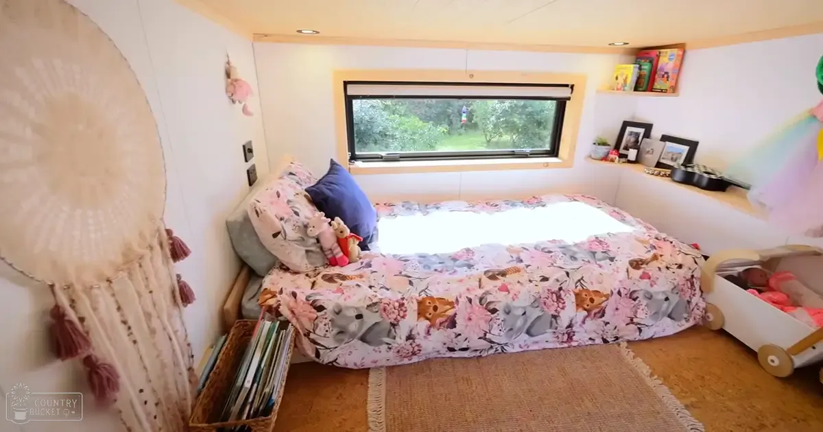 A cozy bedroom scene with floral elephant-patterned bedding, a small window showing greenery outside, bookshelves, a dreamcatcher hanging, and a toy stroller in the corner.