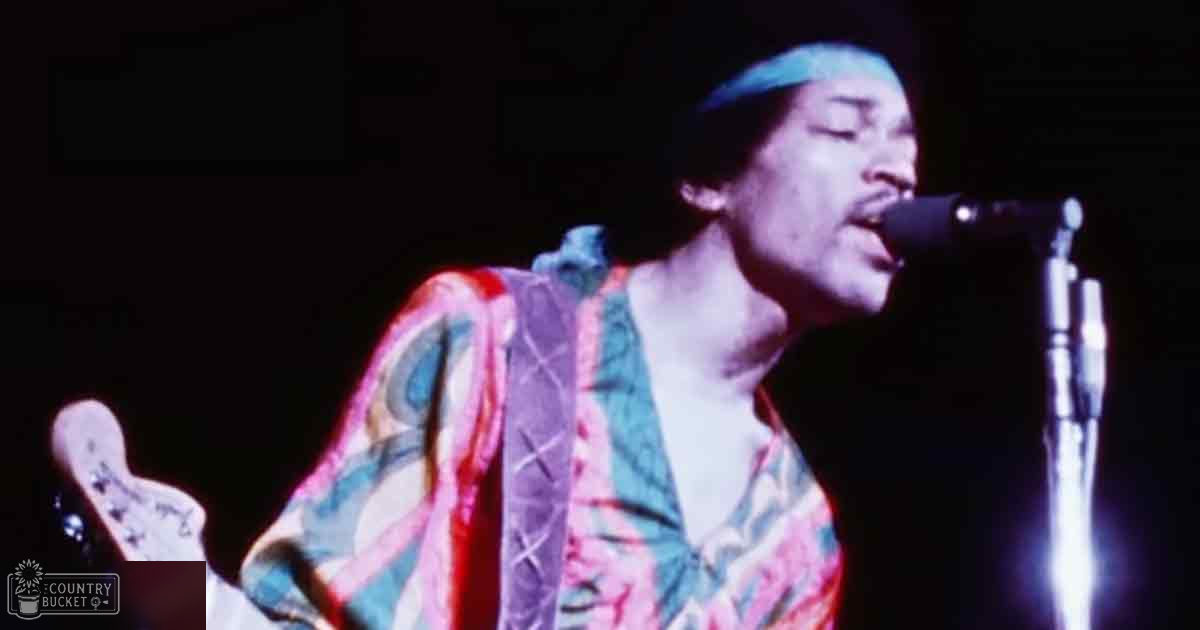 A young adult male singer in a colorful patterned robe on stage during a concert at night, with a microphone stand and guitar visible.