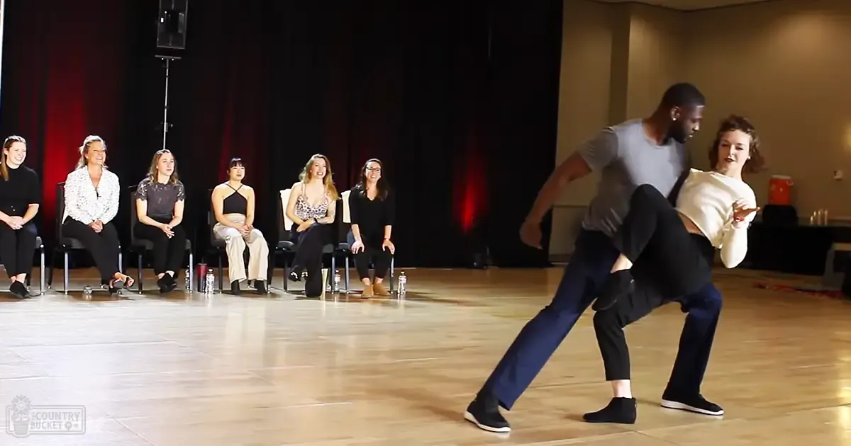 Two adults and six women seated as an audience observe a dance rehearsal taking place indoors with wooden flooring, simple black chairs, and natural light filtering in from one side of the room.