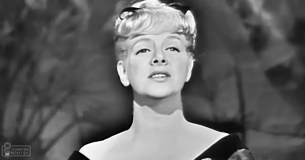 An adult female wearing a form-fitting high-necked dress or blouse stands amidst an outdoor natural setting with trees and branches visible in the background.