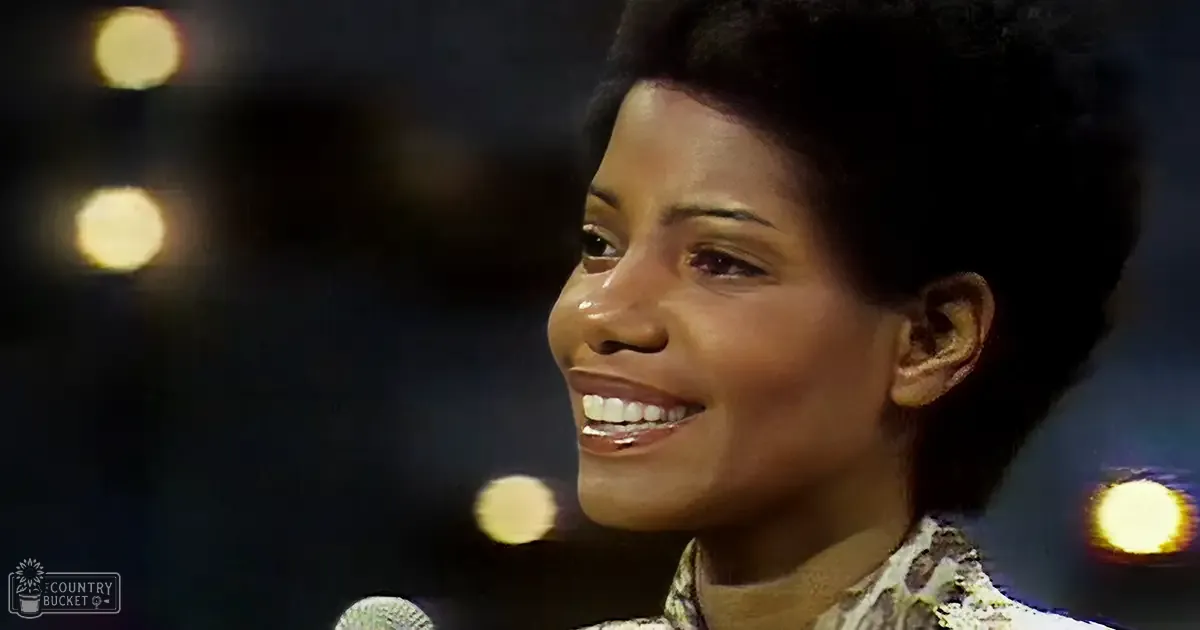 A young adult female wearing a patterned, relaxed-fit blouse with long sleeves, possibly from the 1960s or 70s style, stands on stage under circular bokeh lights, suggesting a concert hall or theater setting. The text 'Madly Odd' in the lower left corner hints at branding.