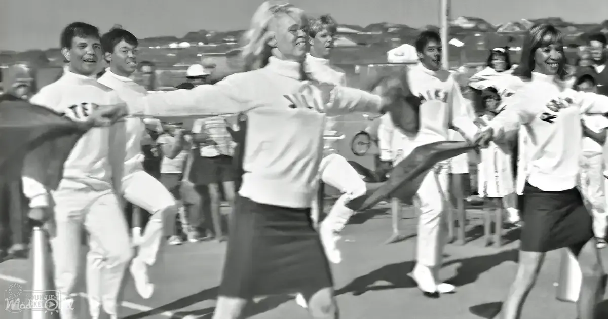 Young adults participating in a dance routine at an outdoor event with spectators on a sunny day.