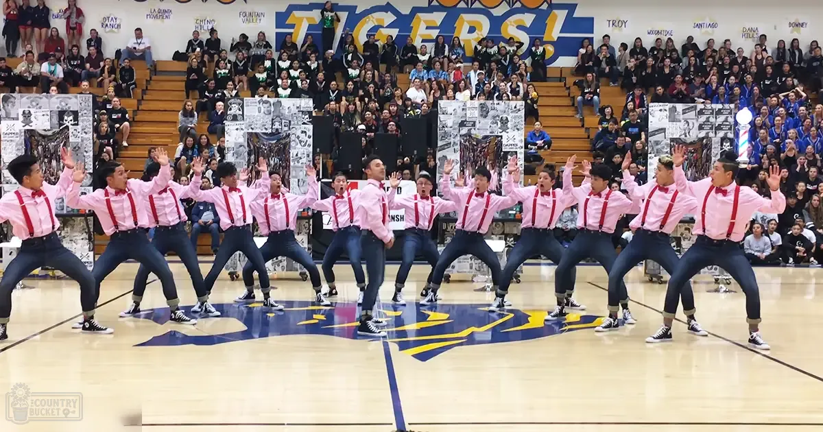 Teenage male dancers performing in pink shirts with red suspenders, black pants, and sneakers at a school event indoors.