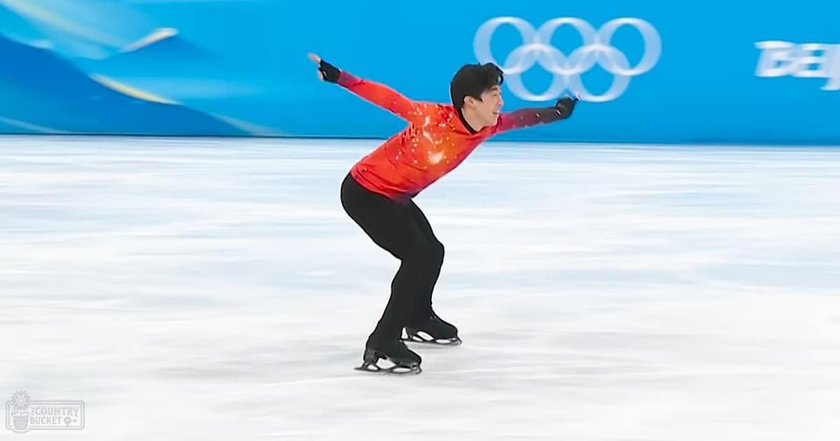 A young adult male figure skater in a red long-sleeve skating costume competes on an indoor ice rink during the Olympic event under bright, even lighting.