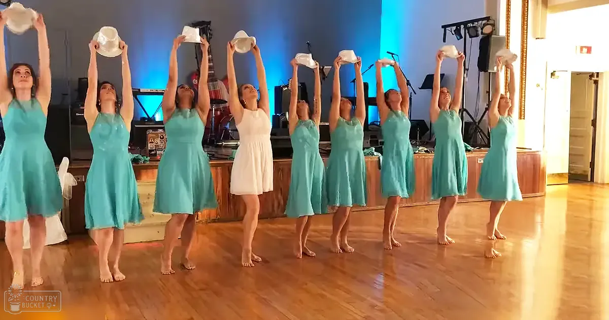 A dance group of young adult females performing on a wooden floor with blue stage lighting, likely an indoor hall or studio at night.
