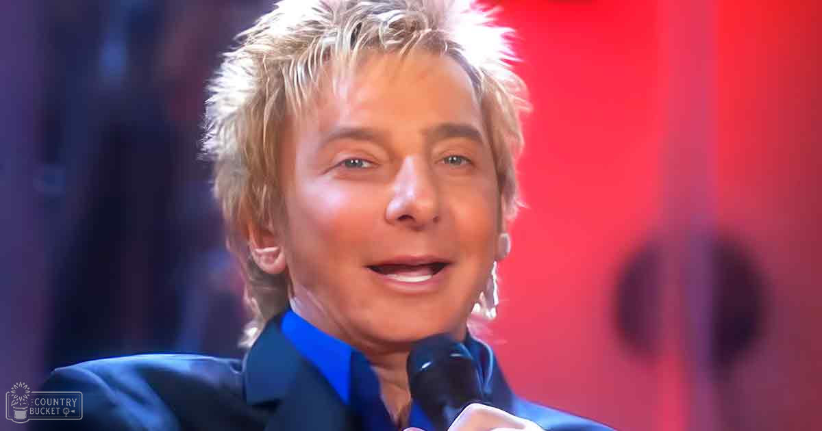 A middle-aged man in a dark blue suit jacket stands on stage, wearing a shiny lapel and red background lights suggest an indoor performance setting.