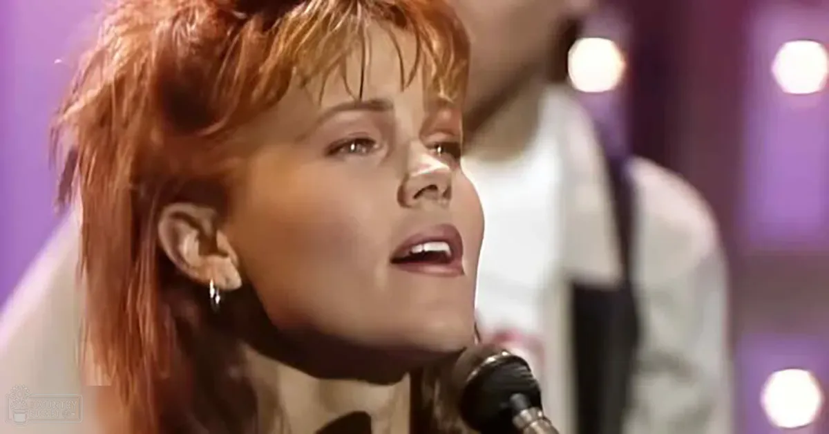 A young adult female singing into a microphone at what looks like a performance setting, wearing a white garment with long sleeves in an indoor venue with purple lighting.