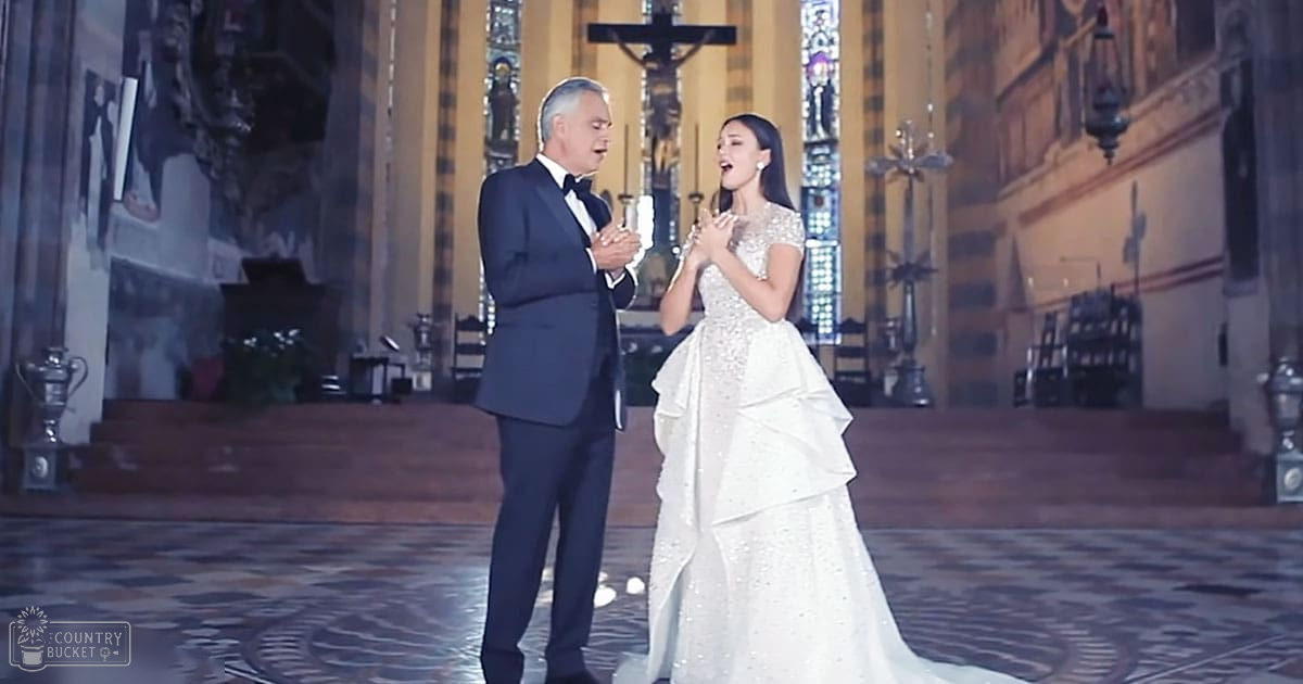 A couple in formal attire stands inside a church with intricate stained glass windows and a crucifix on the altar, suggesting a wedding or special event.