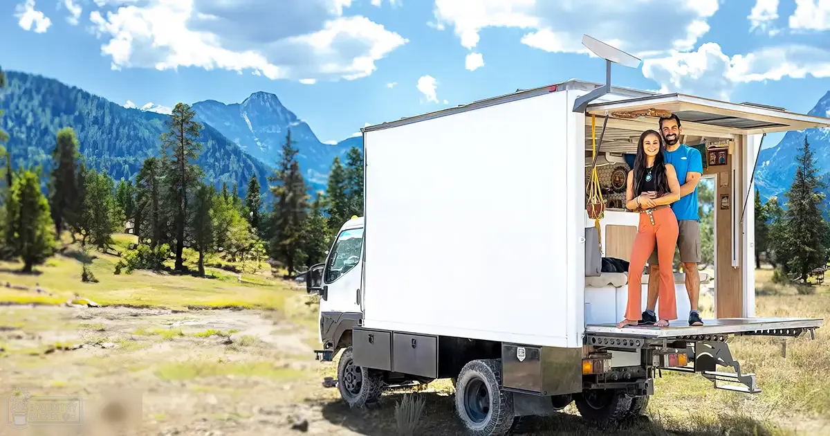 A couple stands in front of their converted truck camper amidst a scenic mountain landscape with lush greenery, clear skies, and outdoor furniture visible inside the camper.